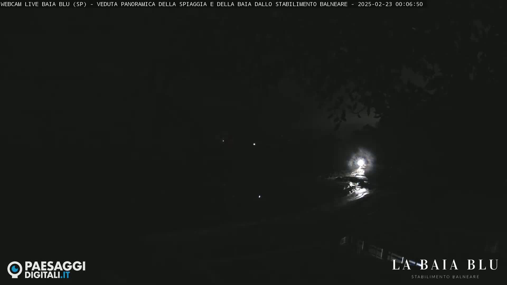 A nighttime, dark scene shows a dimly lit area with some lights visible in the distance, possibly near water.