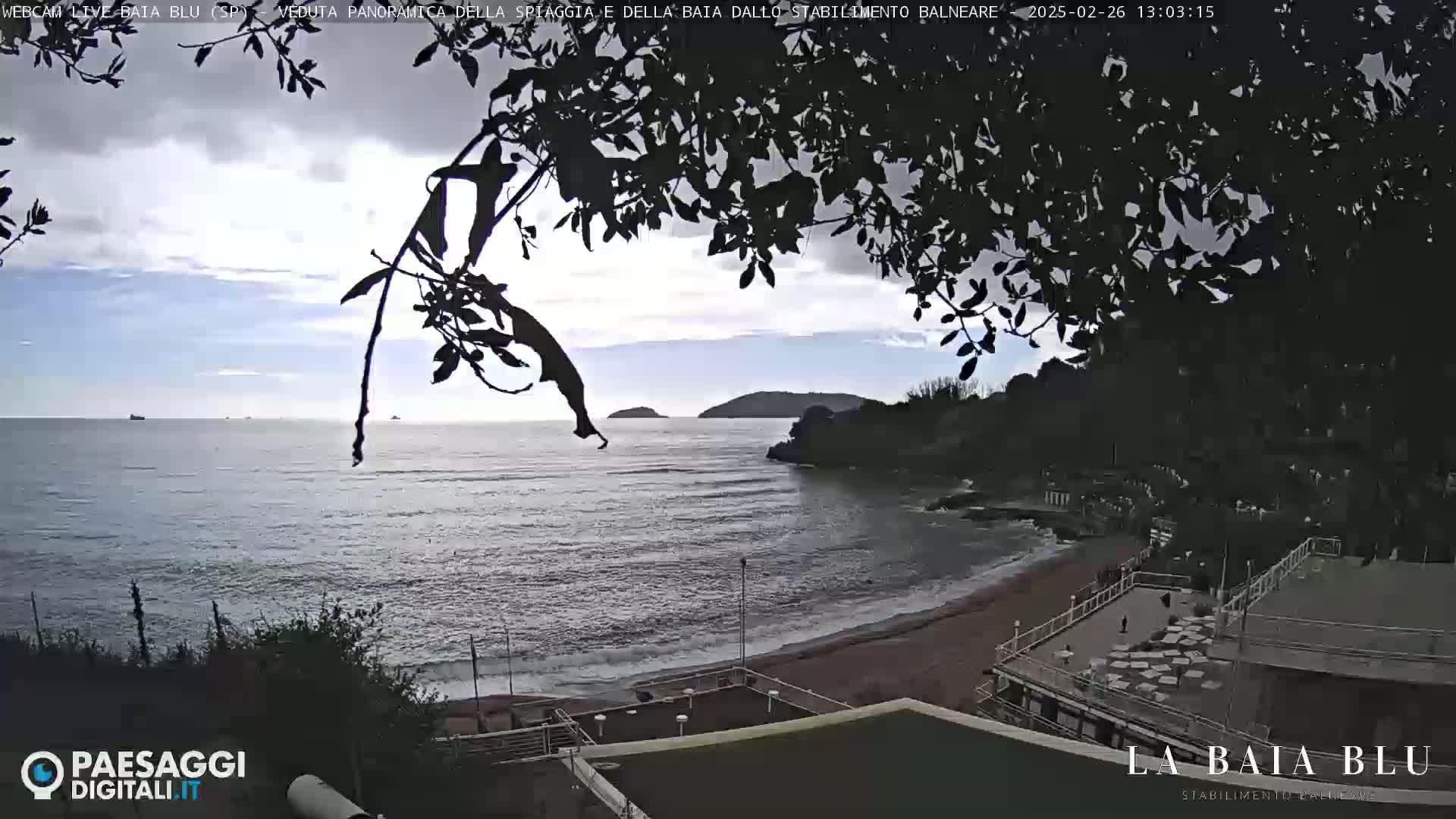 A partly cloudy day overlooking a calm ocean bay with a sandy beach and a beachside building partially obscured by tree branches.