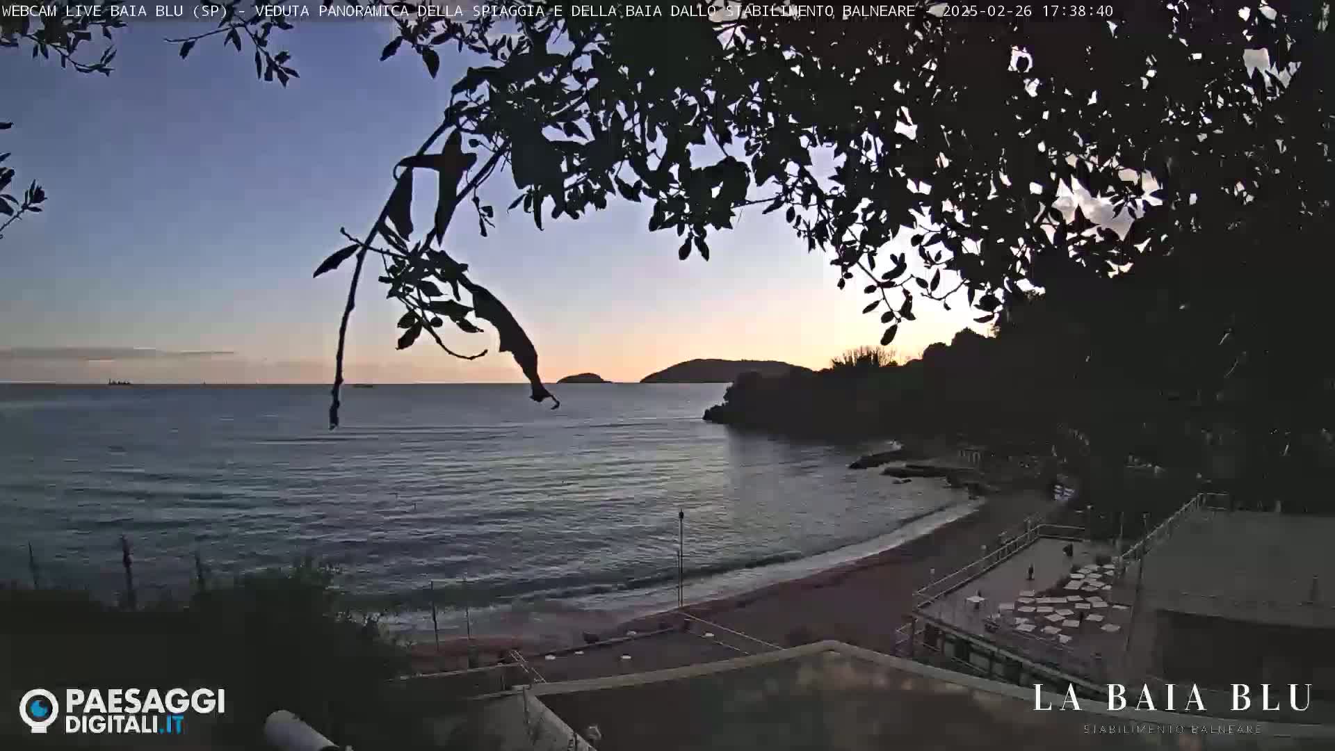 A calm, twilight ocean scene shows a sandy beach with a building at the edge of the water, partially obscured by dark tree branches in the foreground.