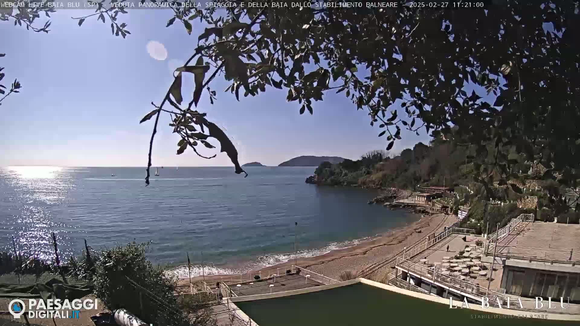 A sunny day overlooking a calm bay with a sandy beach and a beach club, partially obscured by tree branches.