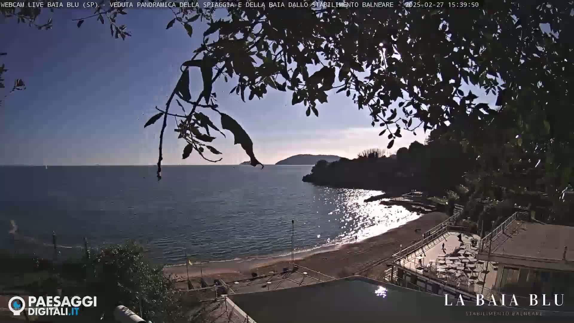 A partly sunny day overlooking a calm bay with a sandy beach and a beach club, partially obscured by tree branches.