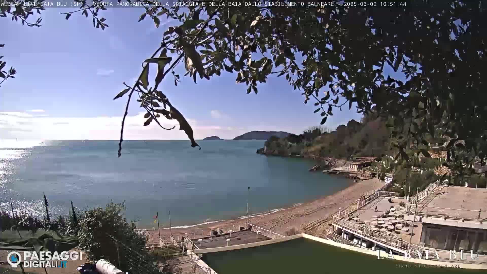 A sandy beach and calm bay are seen under a mostly sunny sky, partially obscured by tree branches in the foreground.