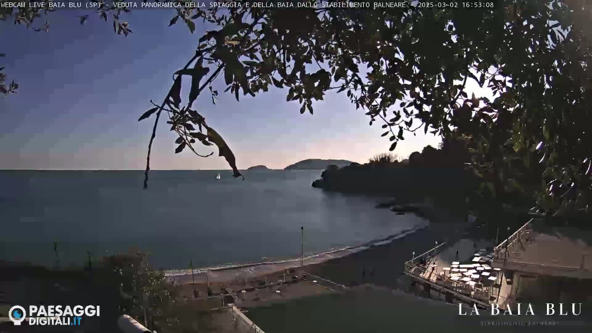 A mostly sunny view of a calm bay with a sandy beach and a building, partially obscured by tree branches.