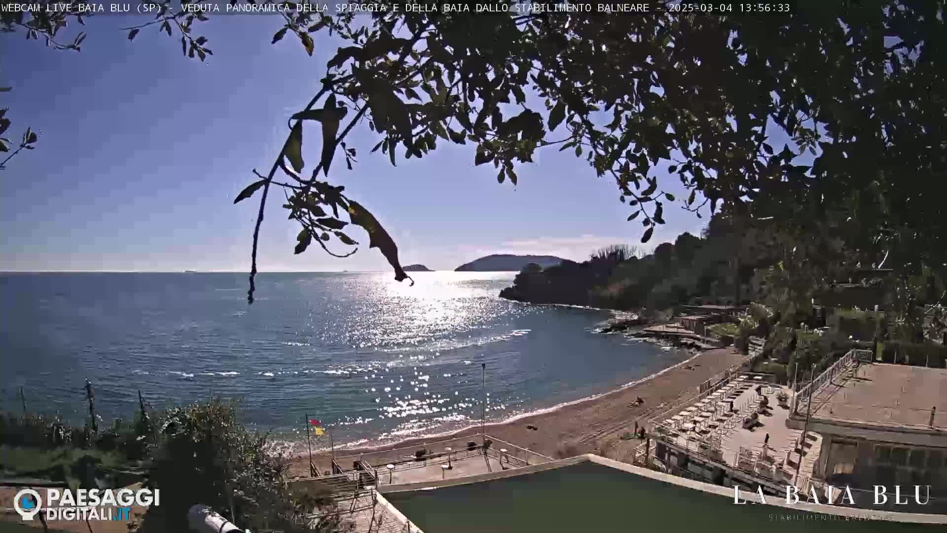A sunny, partly cloudy day overlooks a calm bay with a sandy beach and beach club, partially obscured by tree branches.