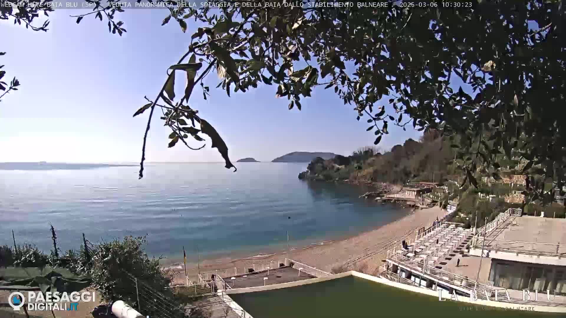 A tranquil beach scene on a sunny day shows a calm bay with a sandy beach and a few buildings nestled amongst greenery, partially obscured by tree branches in the foreground.