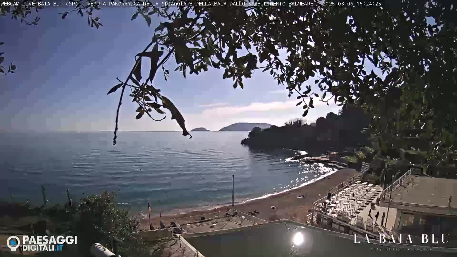 A partly sunny view from a high vantage point shows a calm bay with a sandy beach and a seaside establishment, partially obscured by tree branches in the foreground.