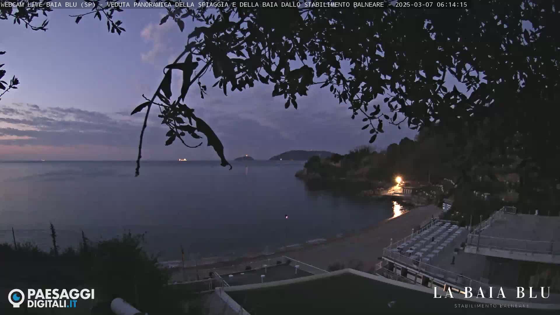A twilight view of a calm bay, partially obscured by silhouetted tree branches, shows a beach with empty seating and a distant shoreline under a partly cloudy sky.