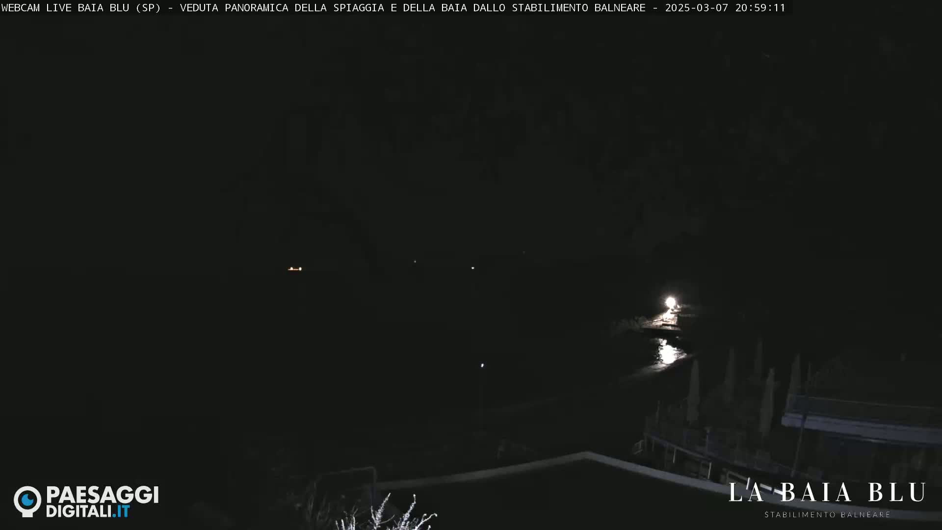 A dark nighttime view of a beach and bay, with a few faint lights visible in the distance.