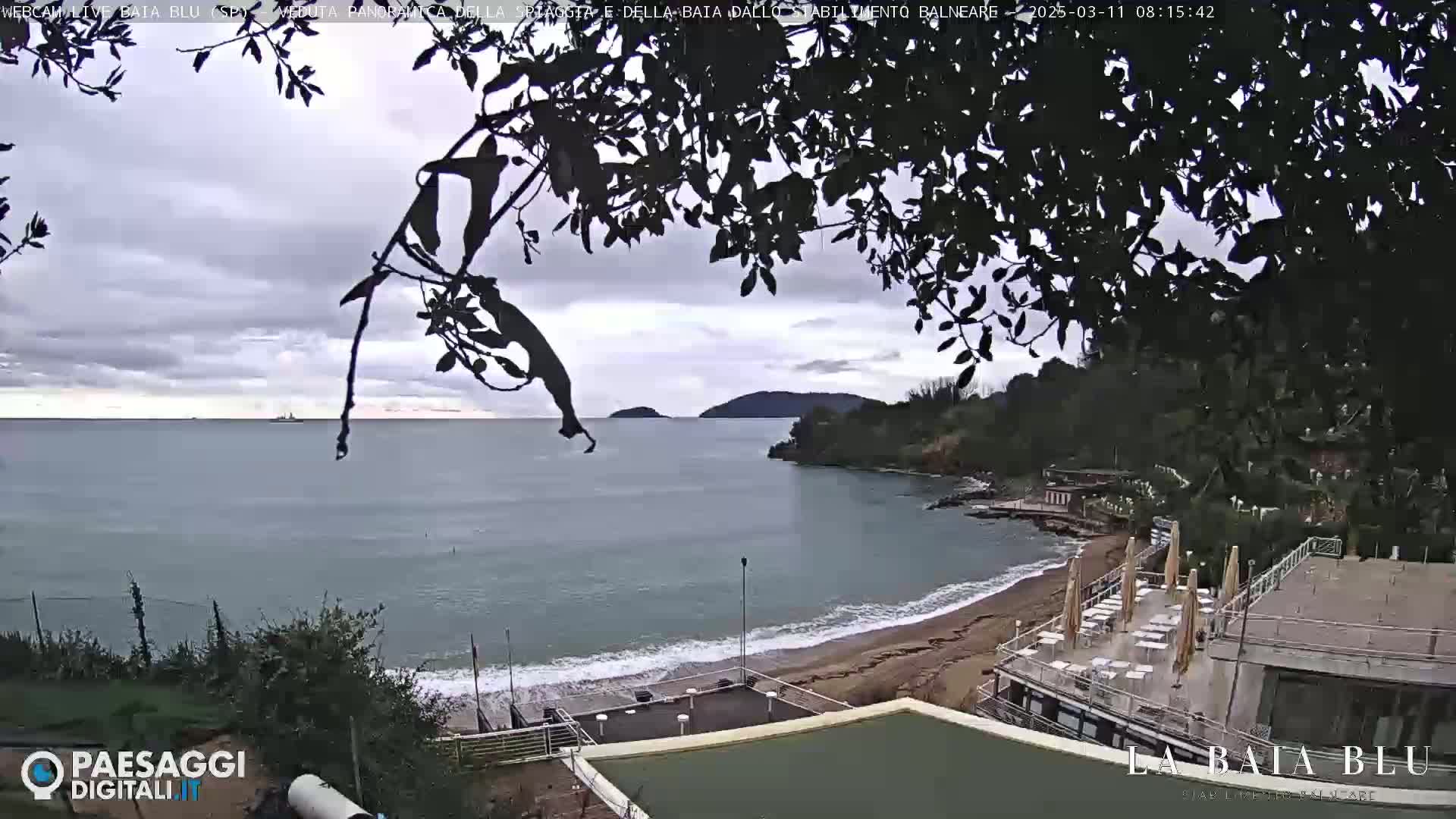 A partially overcast day reveals a sandy beach curving into a calm bay, with buildings and trees visible along the shore and a distant island under a grey sky.