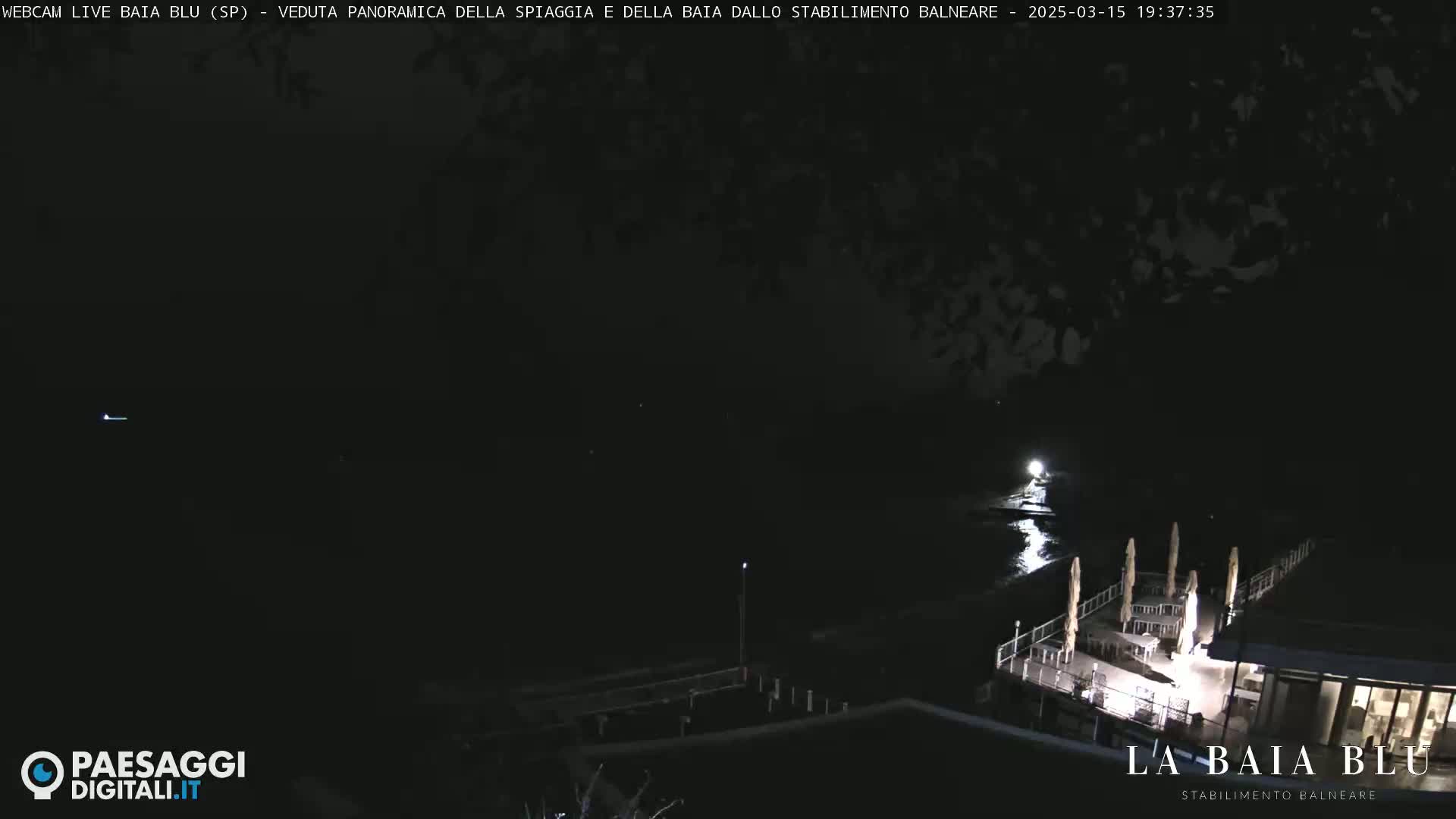 A nighttime, panoramic view of a dark beach and body of water, with a lit outdoor restaurant visible in the foreground.