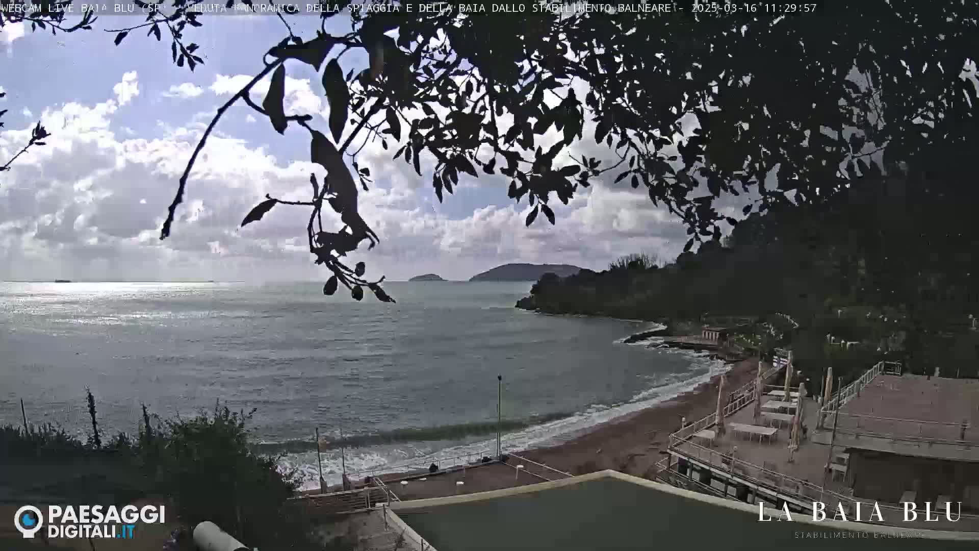 A partly cloudy day at a beach with calm water, viewed from behind tree branches, shows a sandy shore and a building with patio furniture.