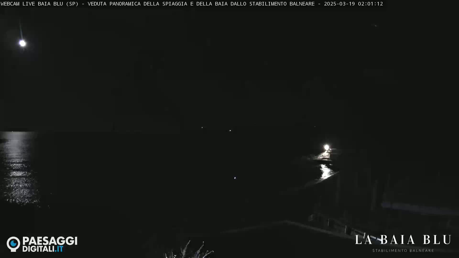 A nighttime view of a dark body of water reflecting a bright moon, with some faint lights visible on the shore.
