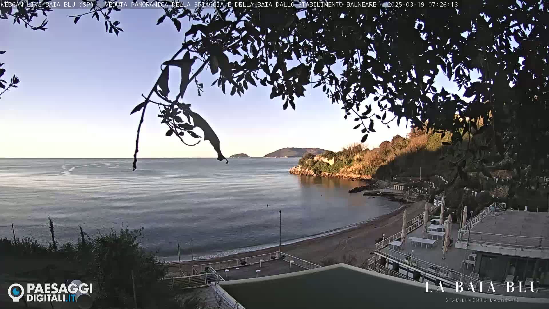 A tranquil coastal scene at dawn, featuring a calm bay with a sandy beach,  overlooked by a building and framed by dark tree branches.