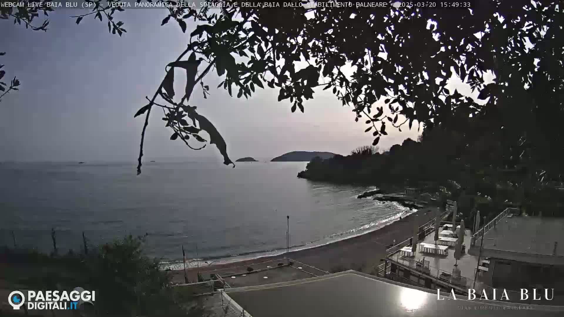 A mostly cloudy day overlooks a calm bay with a sandy beach and some buildings at the shoreline, partially obscured by dark tree branches.