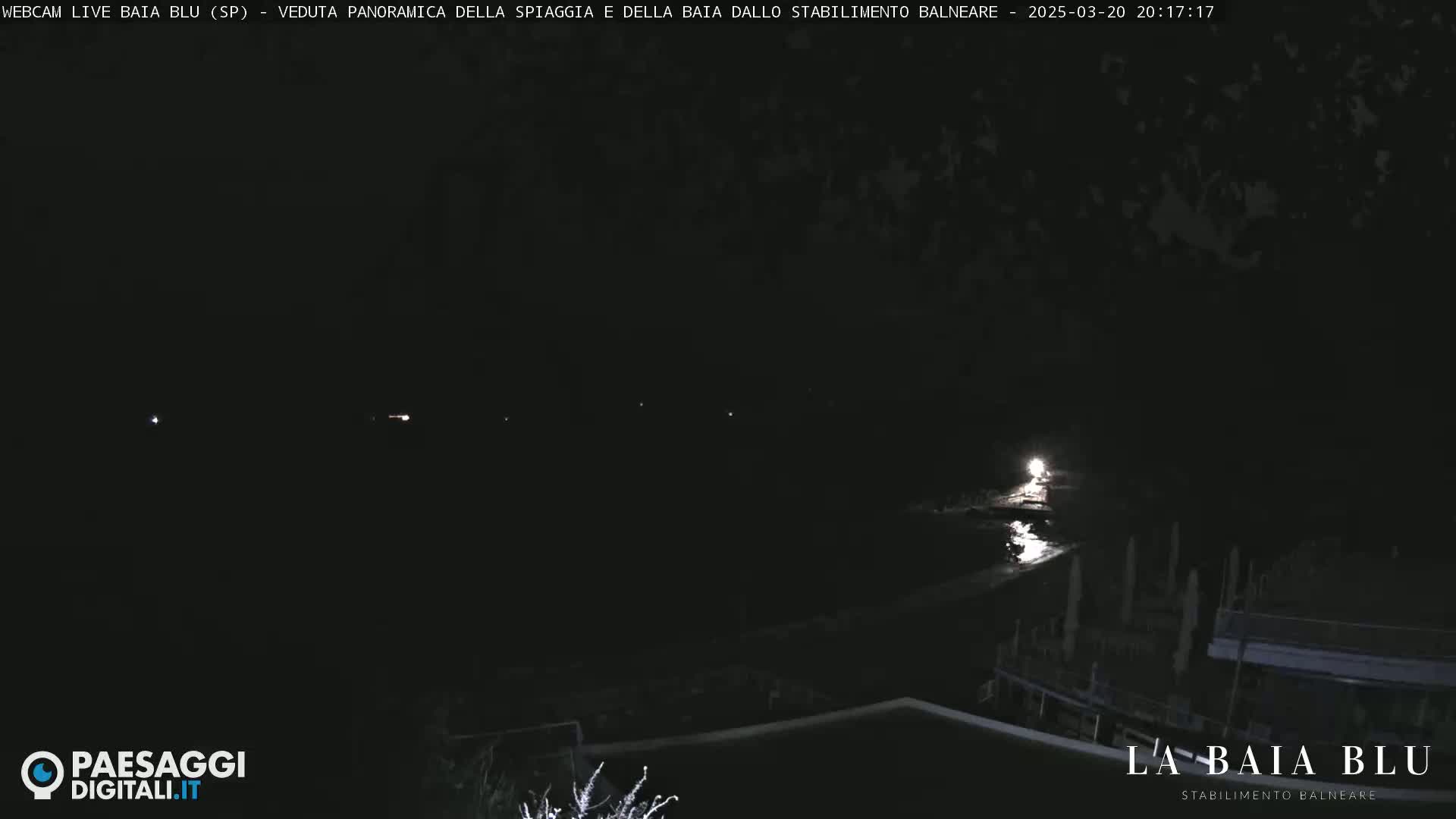 A nighttime, panoramic view of a dark beach and bay with a few faint lights visible in the distance.