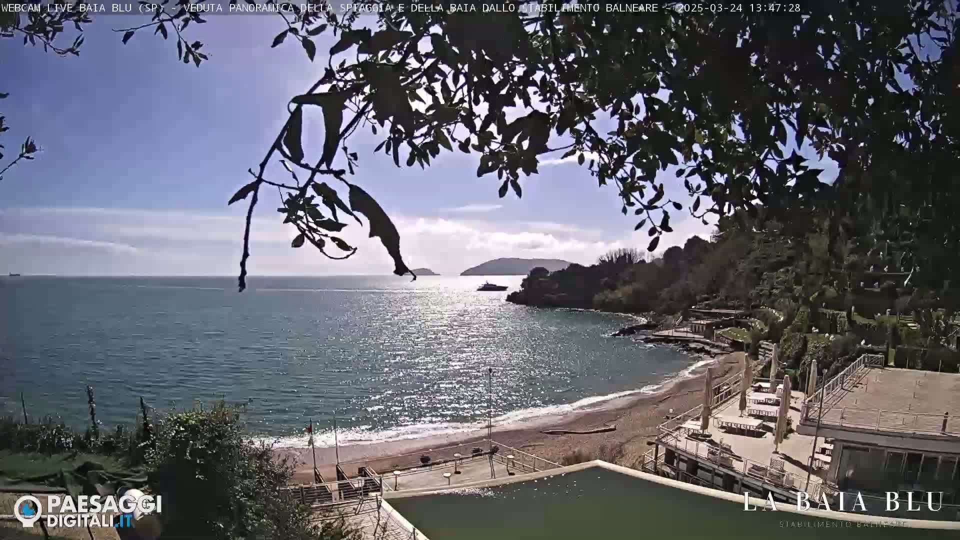 A sunny day at a beach with calm, clear water, viewed from a slightly elevated position showing a sandy beach, buildings, and a boat in the distance.