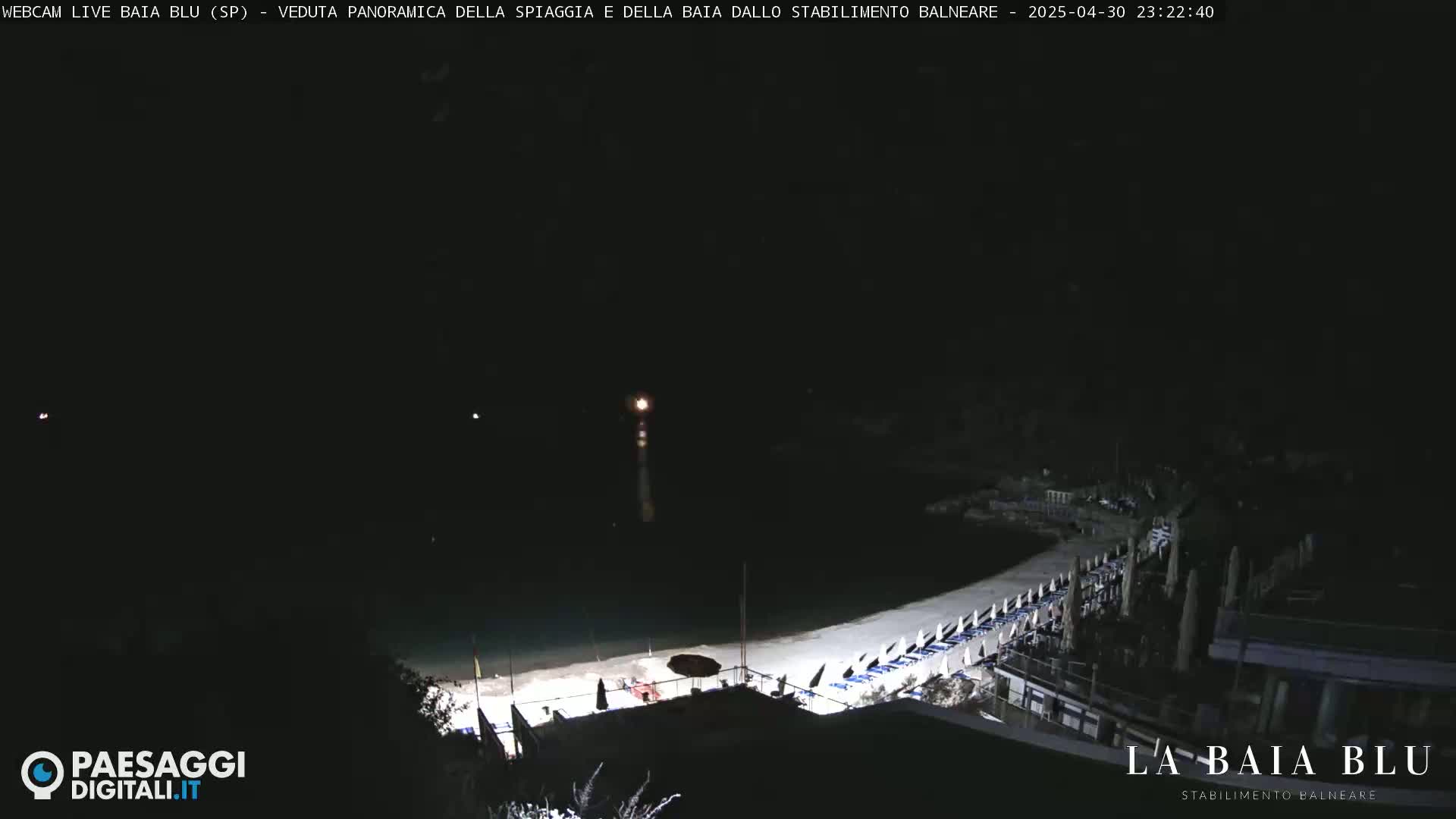A nighttime, panoramic view of a beach with closed umbrellas and chairs, illuminated by artificial light, overlooking a dark body of water.