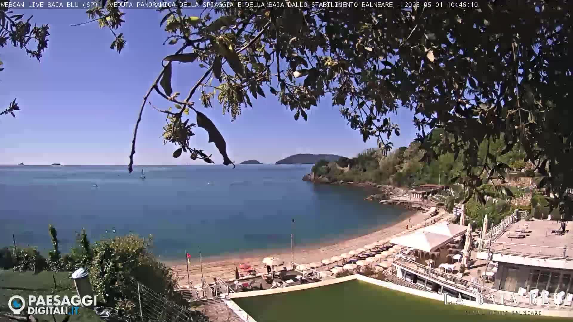 A sunny, clear day overlooking a secluded sandy beach with turquoise water, a beach club, and lush green vegetation.