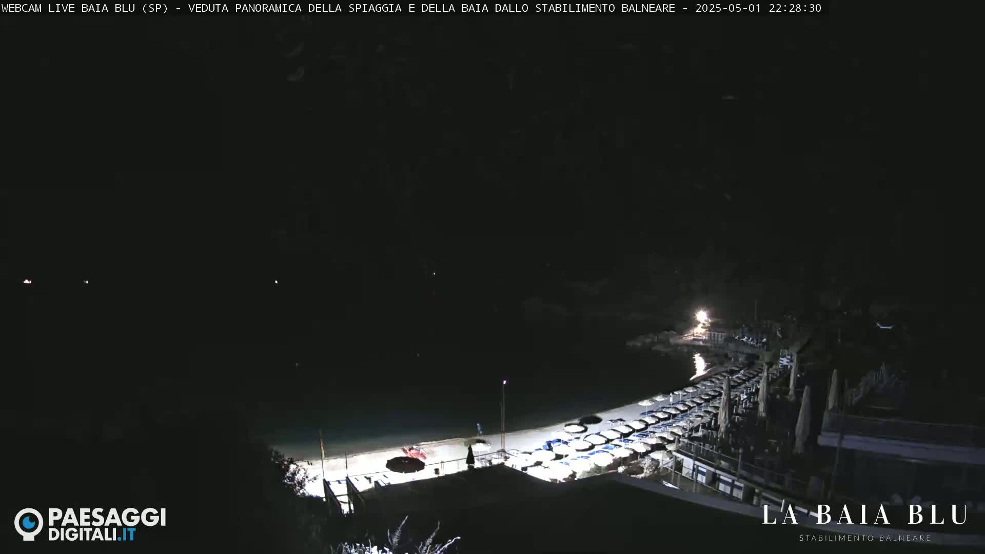 A nighttime, panoramic view of a beach with closed umbrellas and a dark, calm body of water.