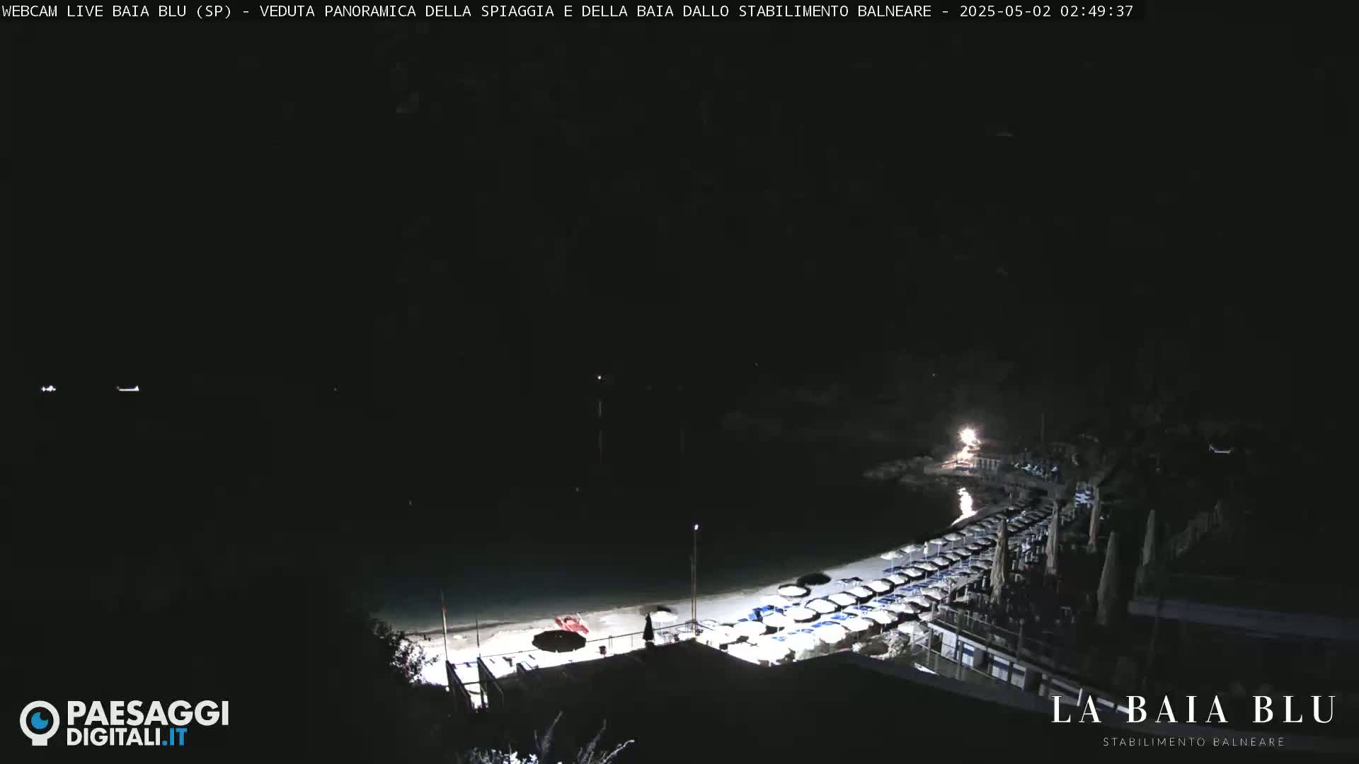 A nighttime, panoramic view of a beach with many closed beach umbrellas and chairs along the shoreline.