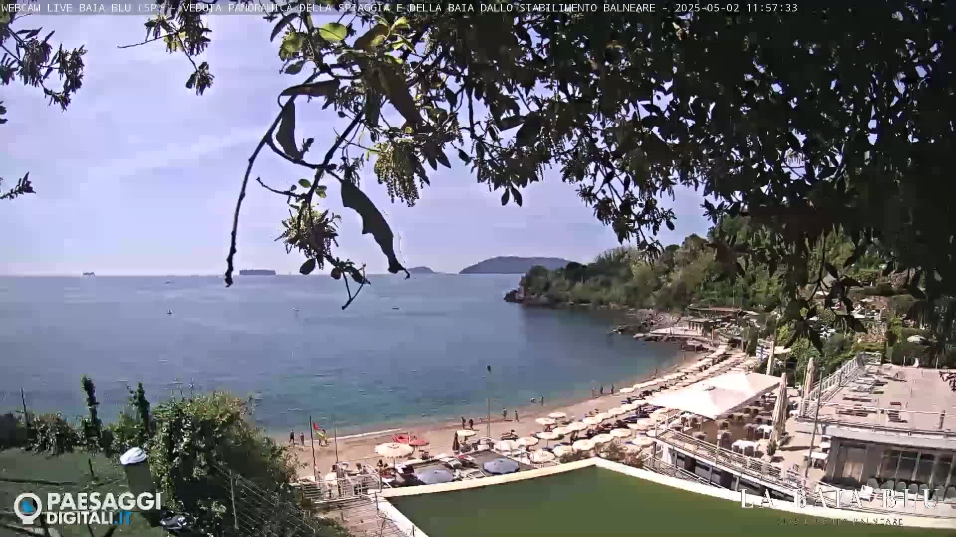 A partly sunny beach scene shows a calm bay with a sandy beach dotted with umbrellas and people, partially obscured by tree branches.