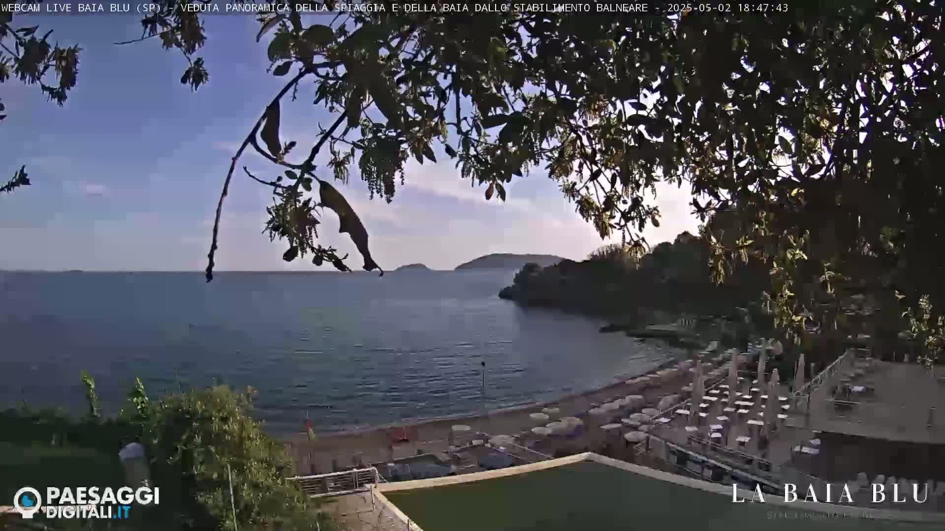 Overlooking a sandy beach with numerous umbrellas and tables, a calm sea stretches towards distant hills under a partly cloudy sky.