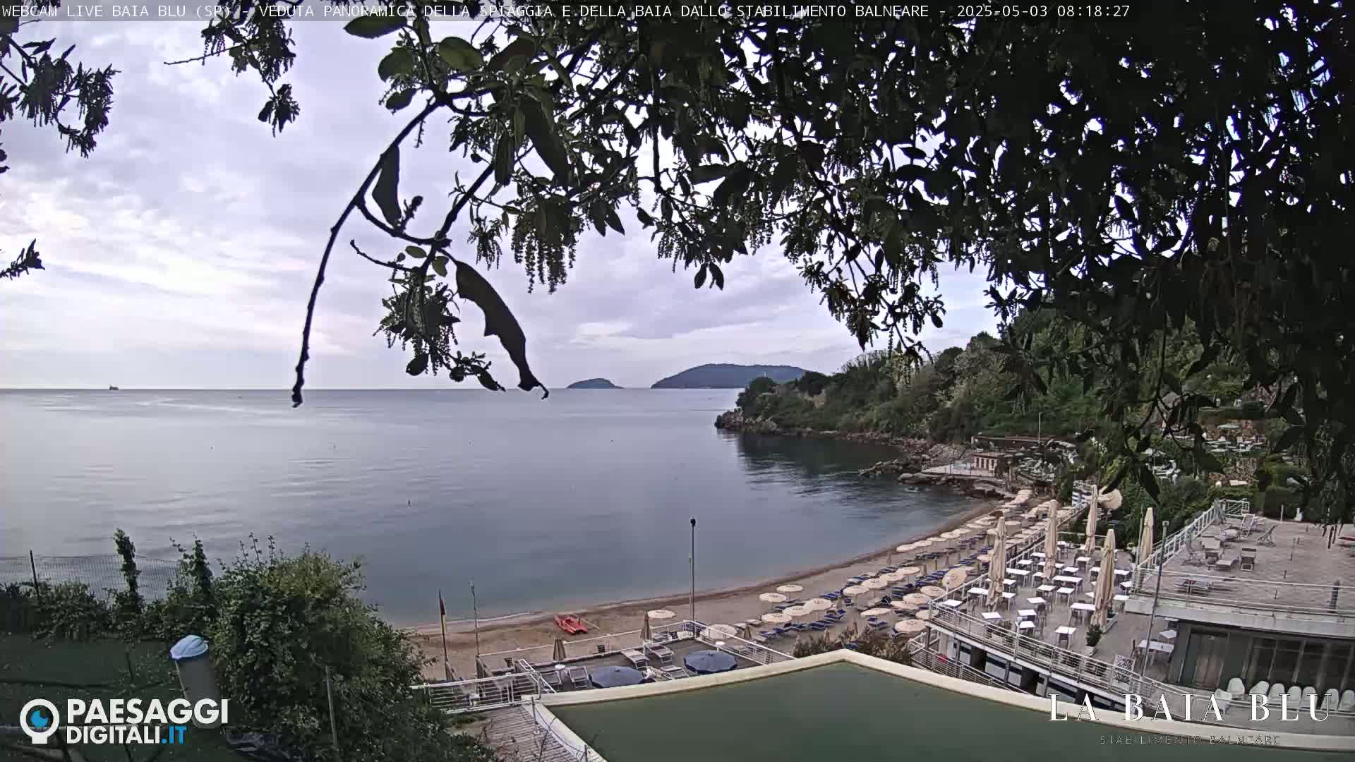 A sandy beach with umbrellas and outdoor seating is nestled in a cove, viewed from above under an overcast sky.