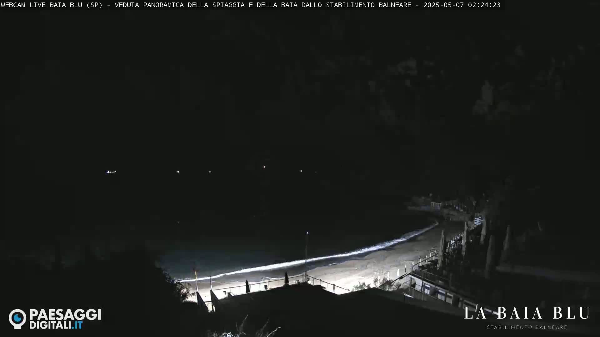 A dark nighttime view of a beach with gently lapping waves, sparsely lit buildings, and a few distant lights.