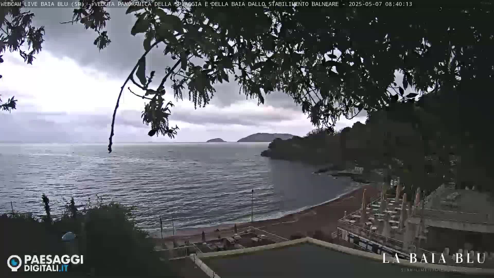 An overcast sky hangs over a calm bay with a reddish-brown sand beach and a shoreline building partially visible through tree branches.