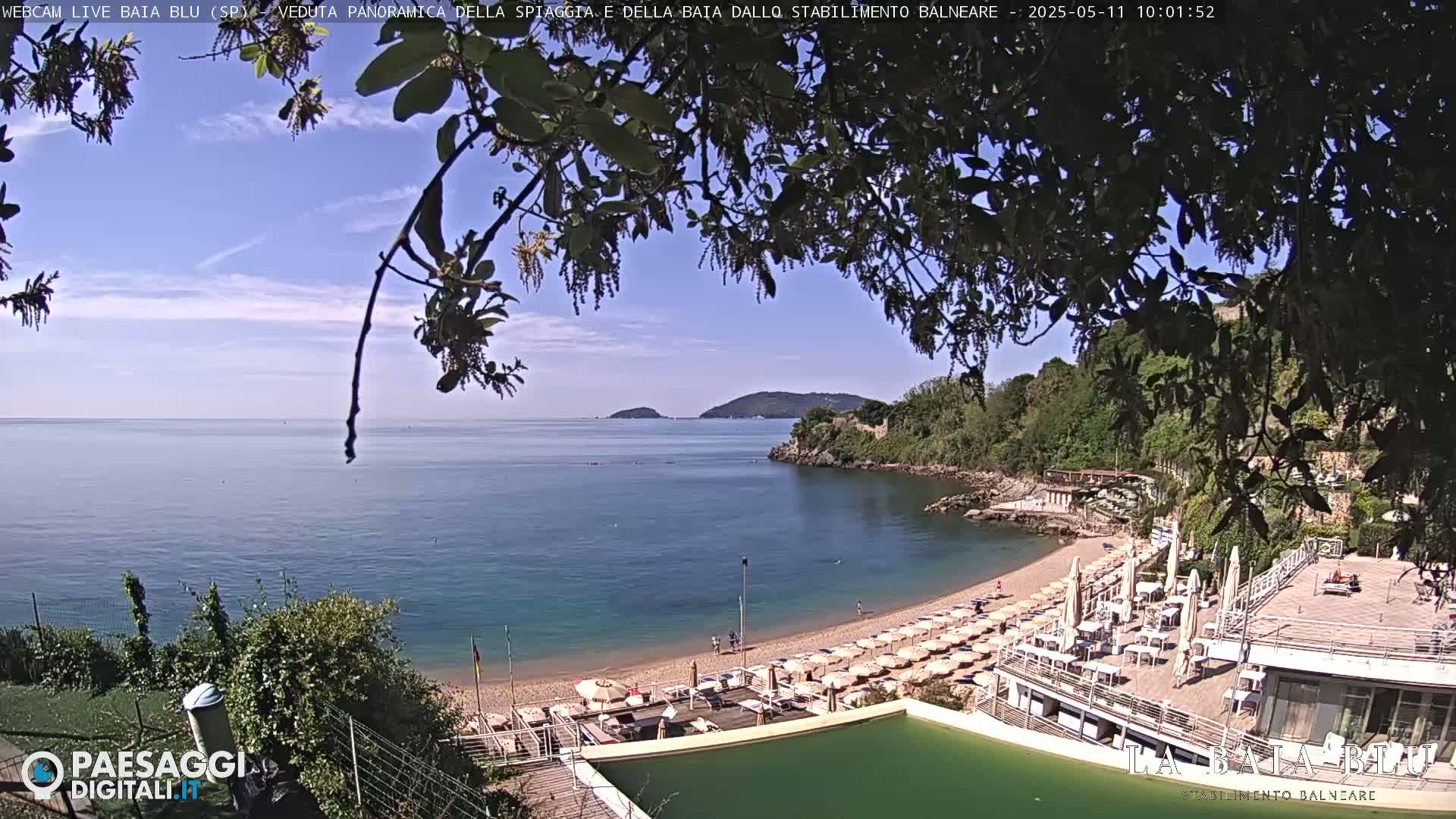 A partly sunny day reveals a sandy beach with numerous umbrellas and lounge chairs at a seaside resort, nestled in a cove with calm blue water and lush green hillsides.