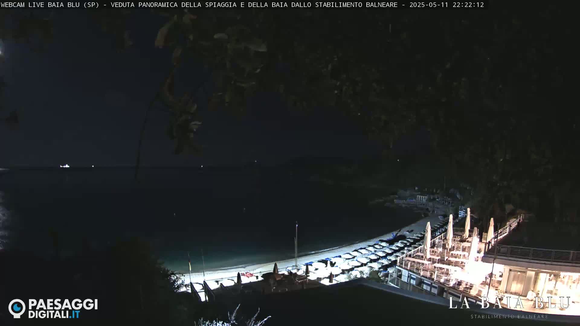 A nighttime view of a beach with numerous closed umbrellas and a lit-up beachfront restaurant overlooking a dark, calm sea.