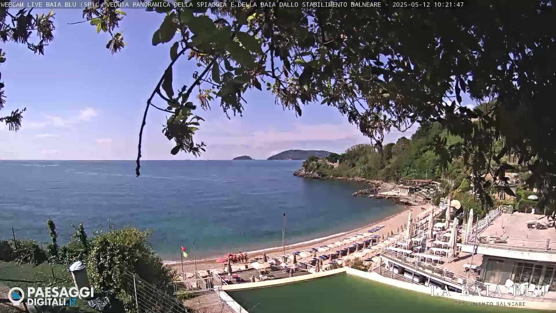 A partly sunny day overlooking a calm bay with a sandy beach, lined with umbrellas and lounge chairs, next to a resort with a pool.