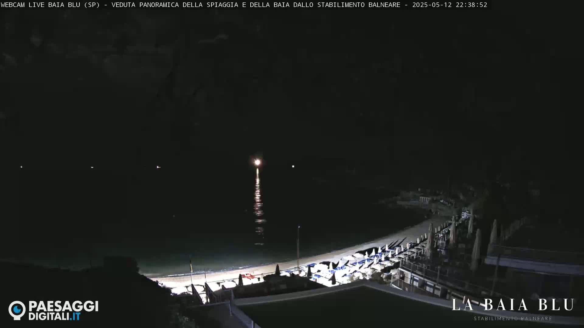 A nighttime, panoramic view of a beach with rows of closed beach umbrellas and chairs, illuminated by faint lights, overlooking a dark body of water.