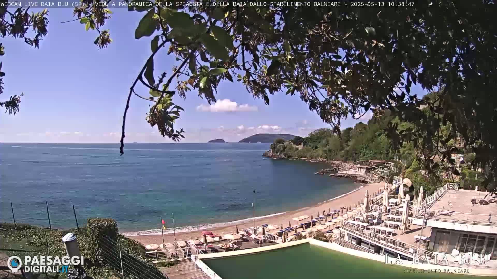 A sandy beach with umbrellas and lounge chairs is nestled in a cove, overlooking a calm blue sea and distant islands under a partly cloudy sky.
