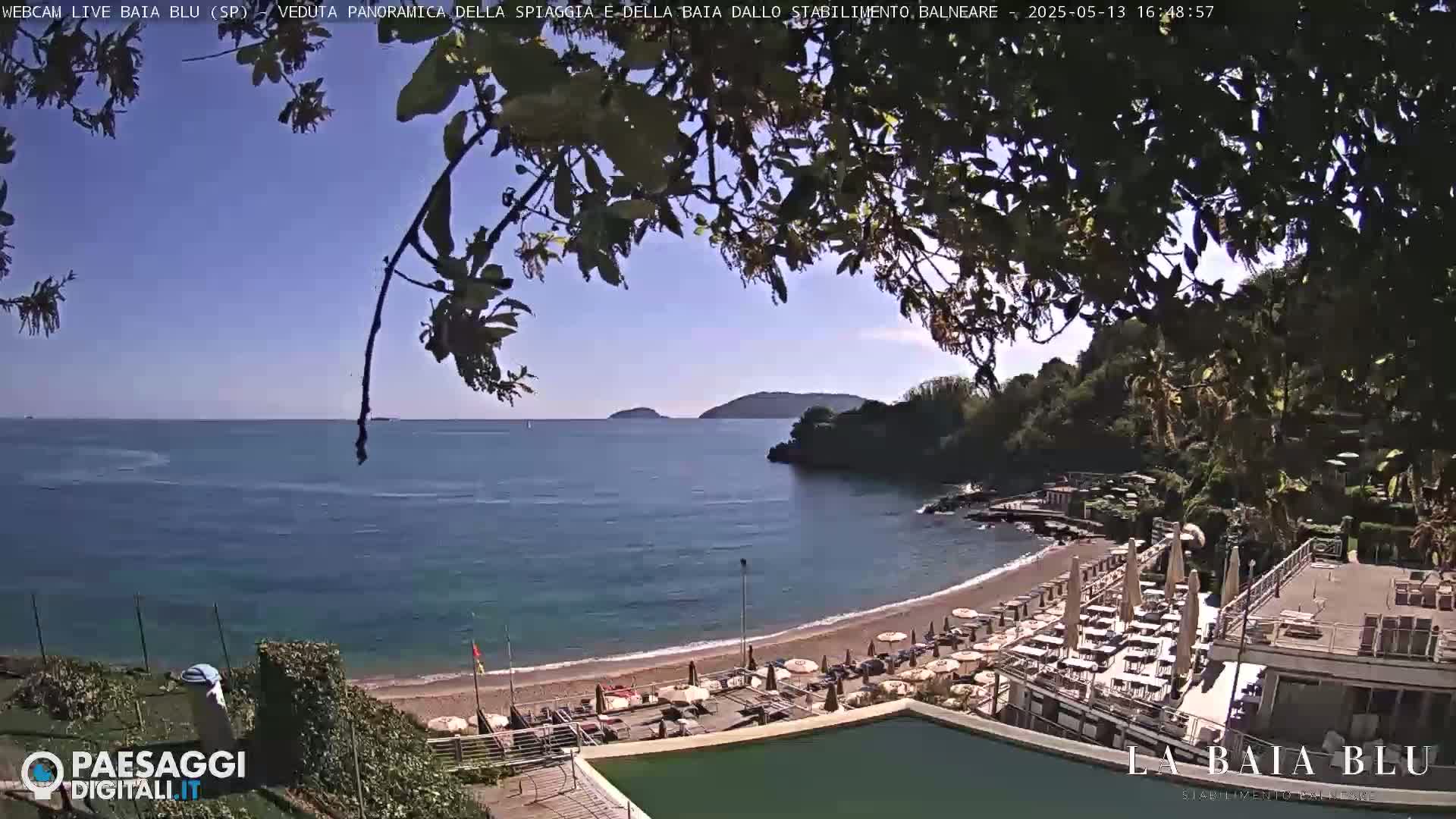 A sunny day reveals a sandy beach with umbrellas and lounge chairs, nestled in a cove next to calm blue water,  with hills and greenery in the background.