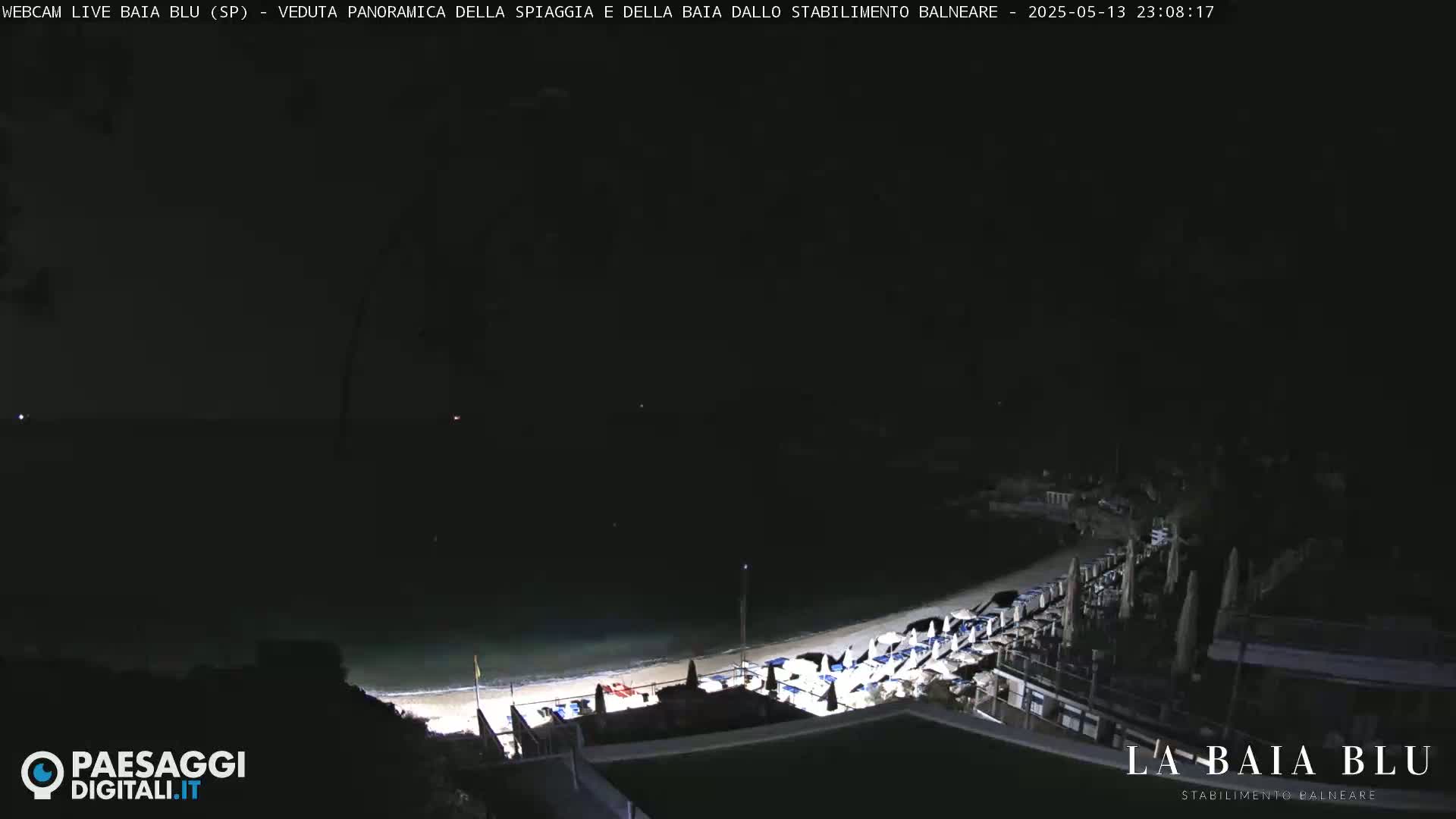 A nighttime, panoramic view of a beach with closed beach umbrellas and chairs, and dark water.