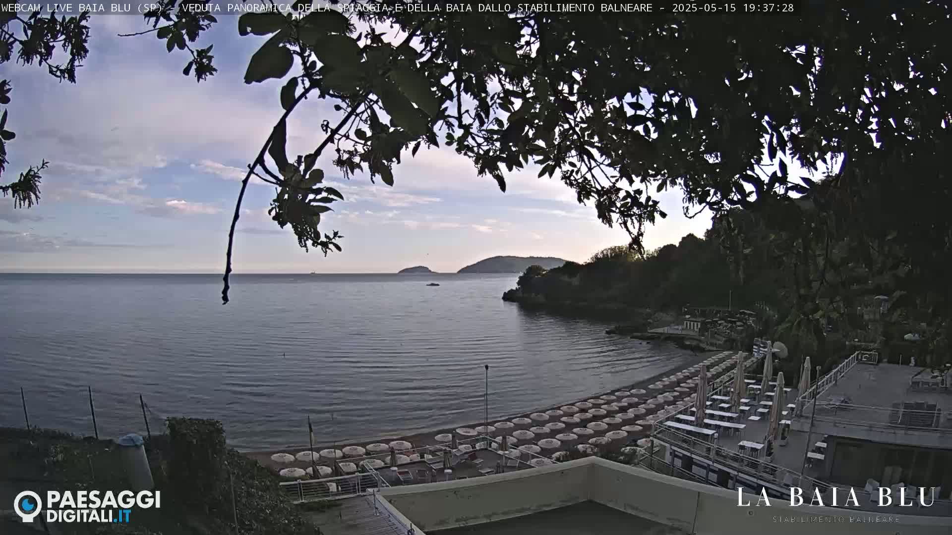 A partly cloudy evening view of a beach with numerous closed umbrellas and outdoor seating areas, overlooking a calm sea and distant hills.