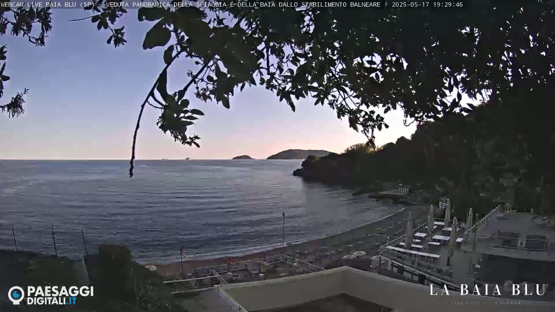 A tranquil beach scene at dusk shows a calm sea, a sandy shore, and a seaside establishment partially obscured by tree branches.