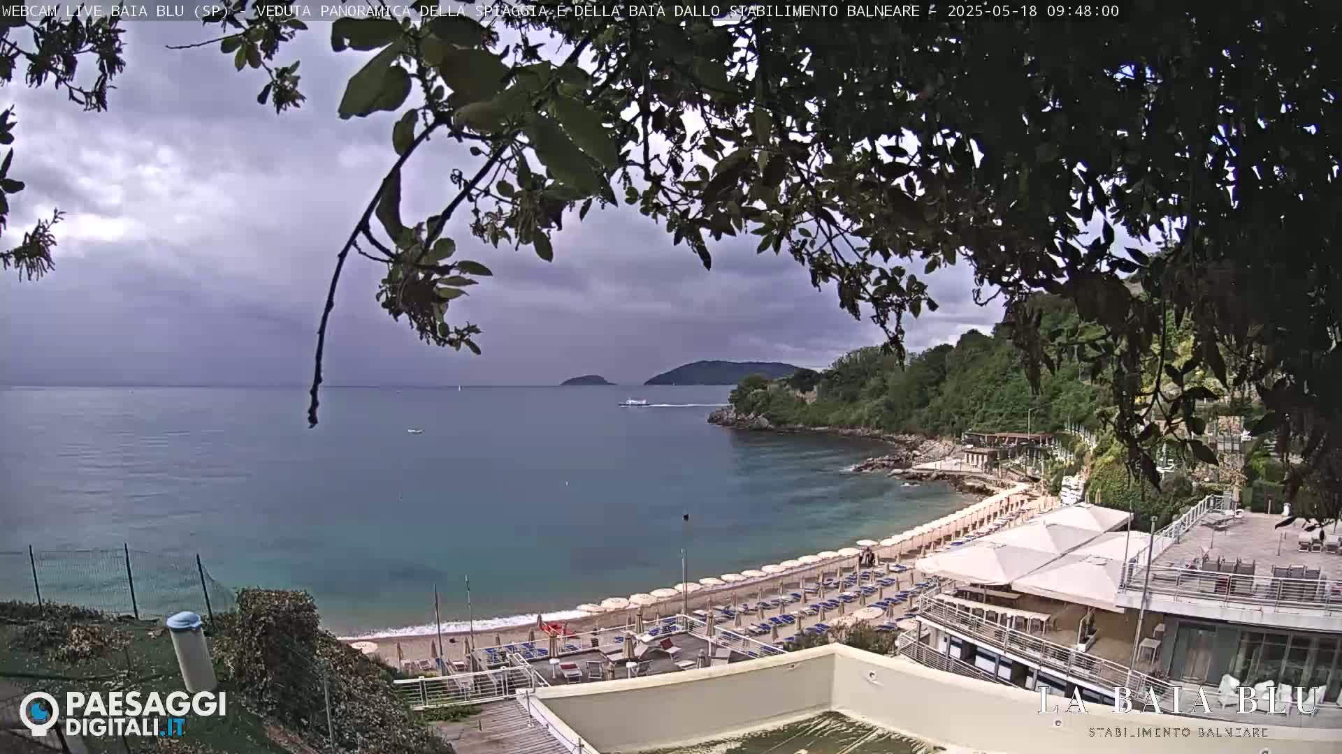 A partly cloudy day overlooks a sandy beach with numerous umbrellas and lounge chairs, a calm teal bay, and green hills in the background.