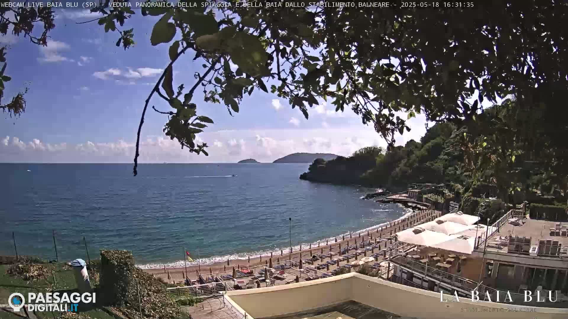 A partly sunny day at a beach with calm blue water, a sandy shore lined with beach umbrellas and lounge chairs, and a backdrop of green hills.