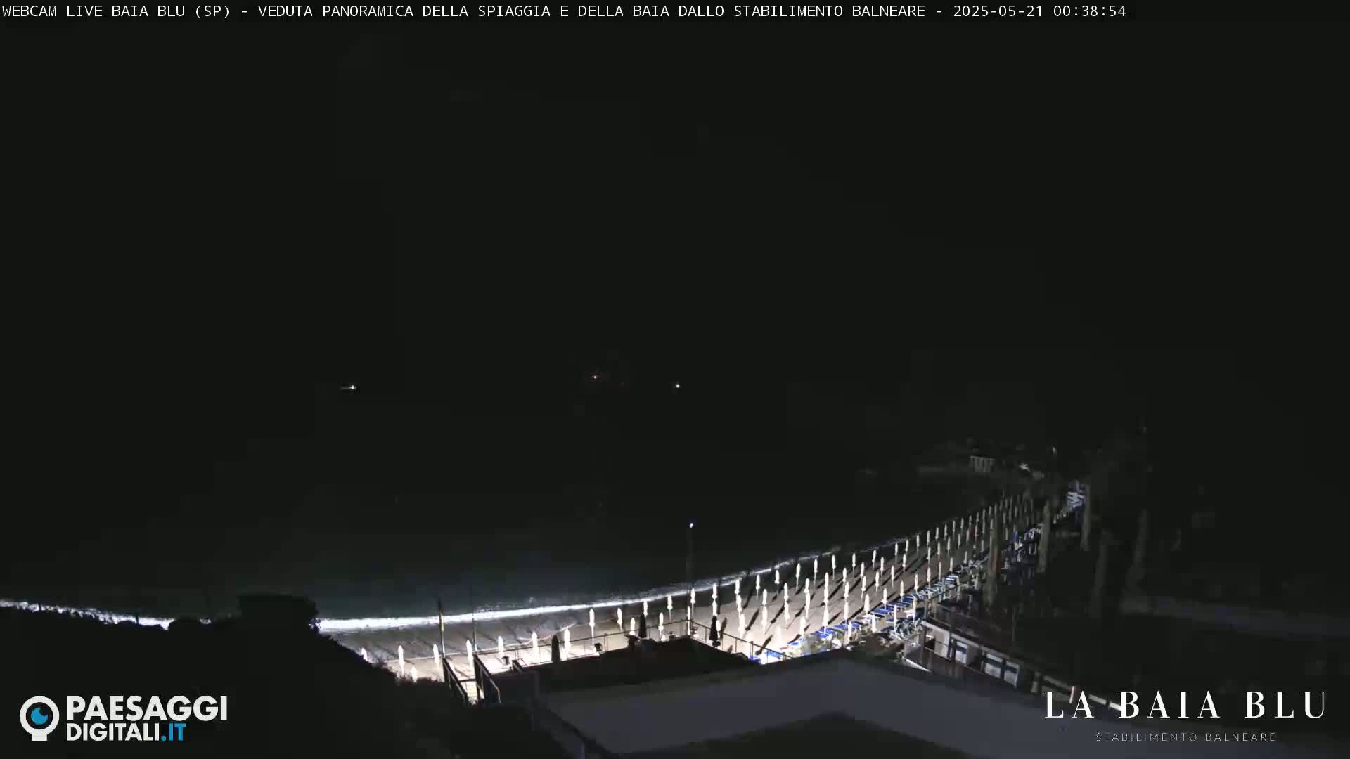 A nighttime, panoramic view of a beach with closed umbrellas and a dark ocean with small waves.