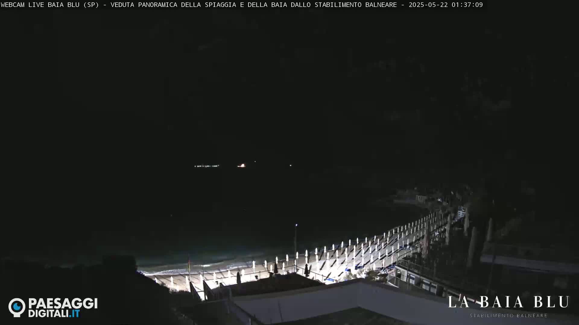 A nighttime, panoramic view of a beach with closed umbrellas and a boardwalk extending into the dark ocean.