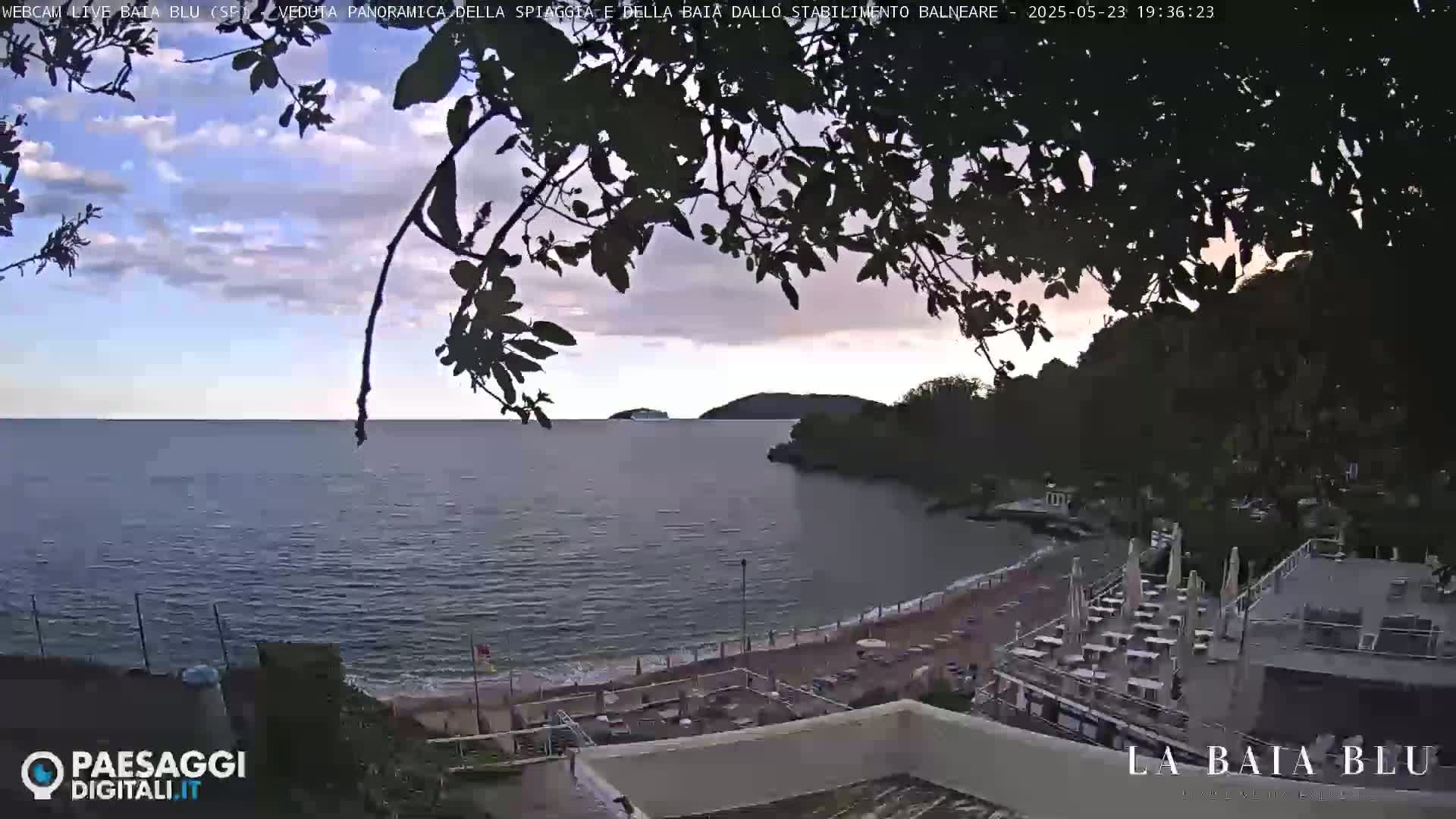 A partly cloudy evening scene overlooks a calm bay with a sandy beach and outdoor seating area.