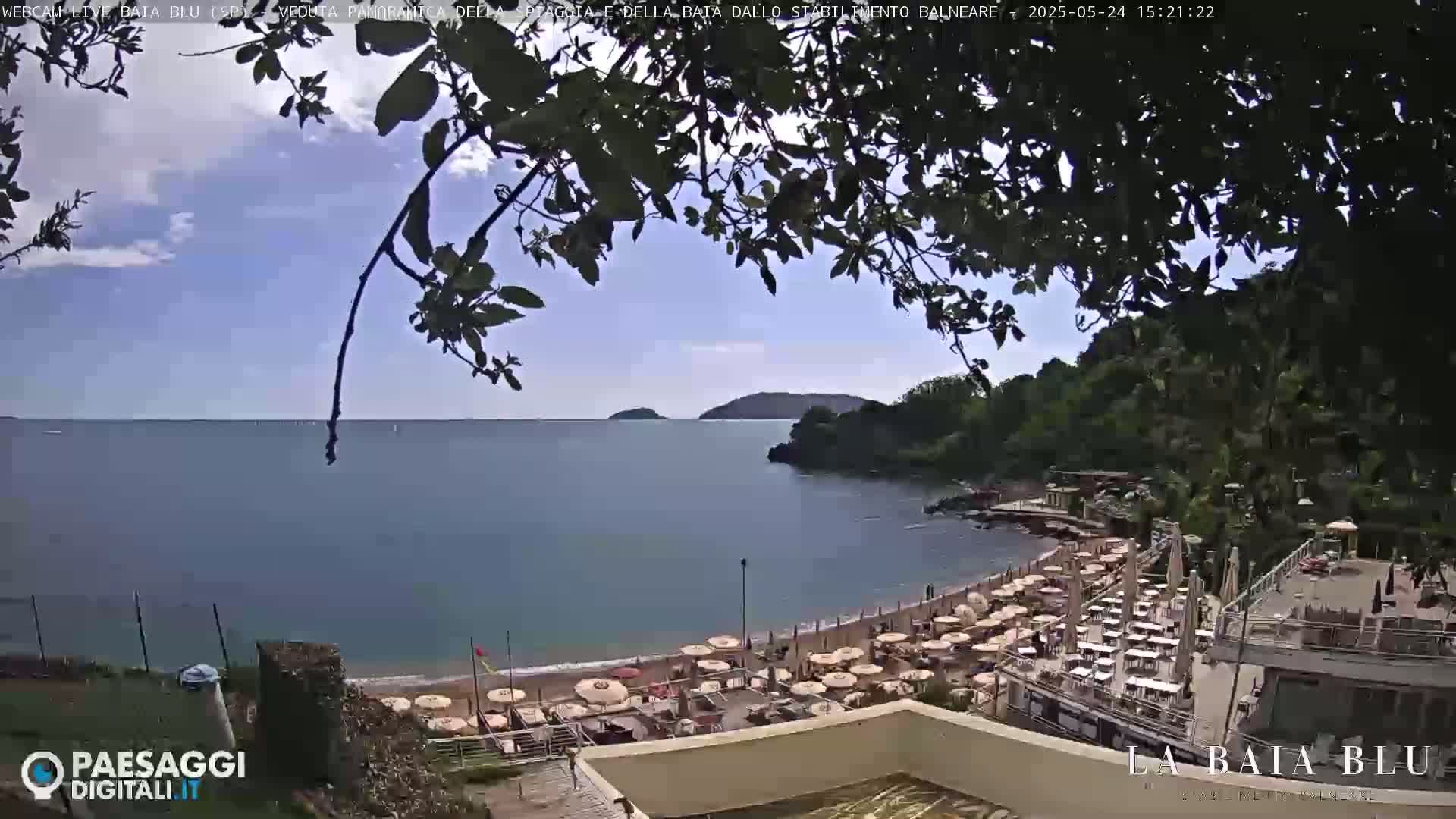 A partly sunny day overlooks a calm bay with a sandy beach lined with umbrellas and beach chairs, partially obscured by tree branches.