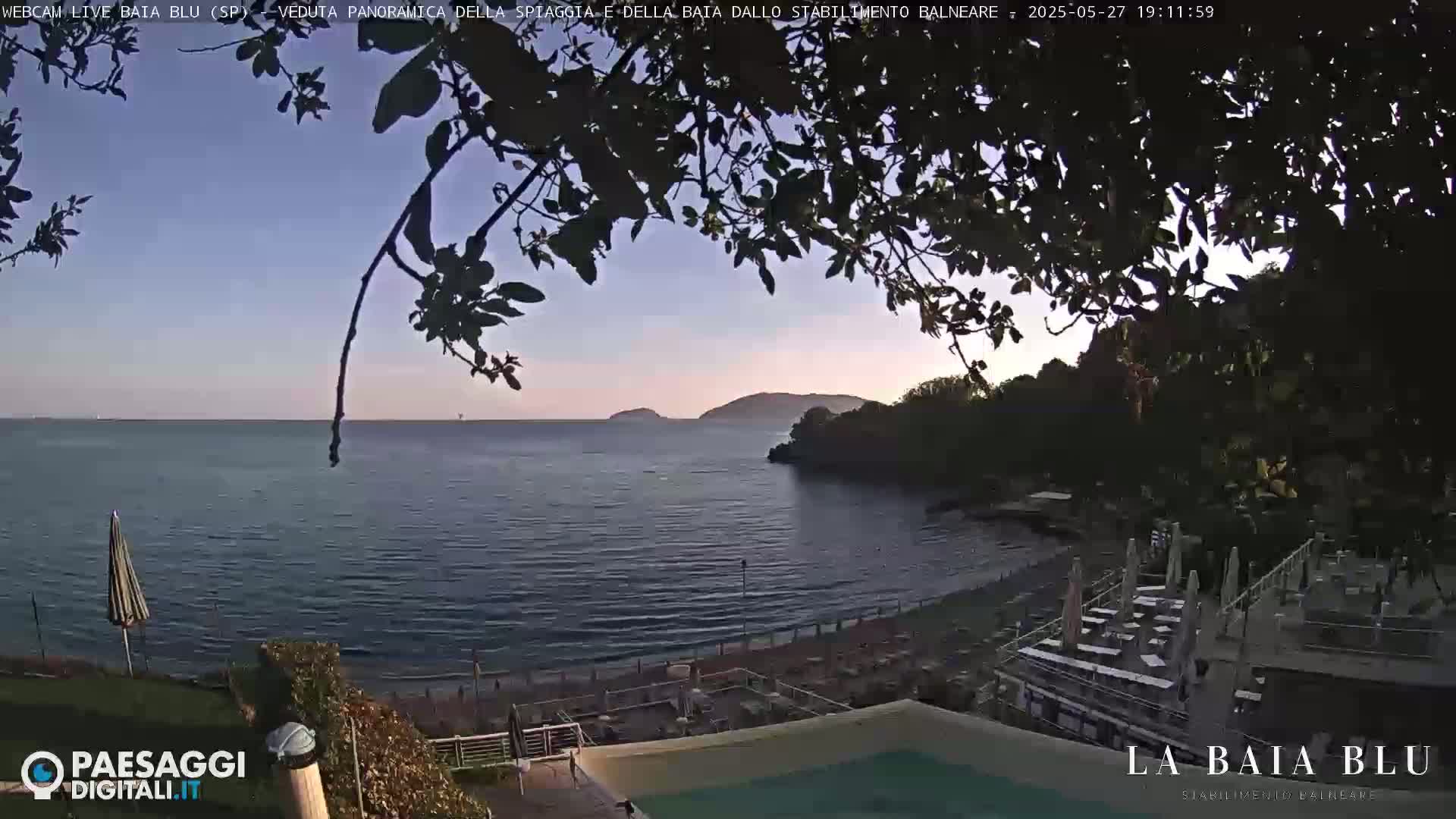 A calm, twilight ocean scene is viewed from behind leafy branches, showing a beach with patio furniture and a swimming pool at a resort, with islands visible in the distance.