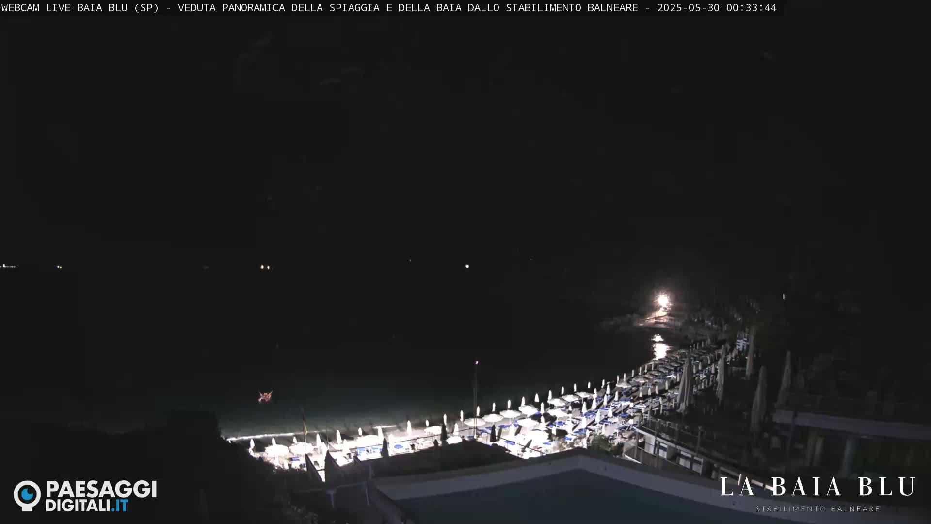 A nighttime, panoramic view of a beach with rows of closed umbrellas and dimly lit chairs, overlooking a dark body of water.