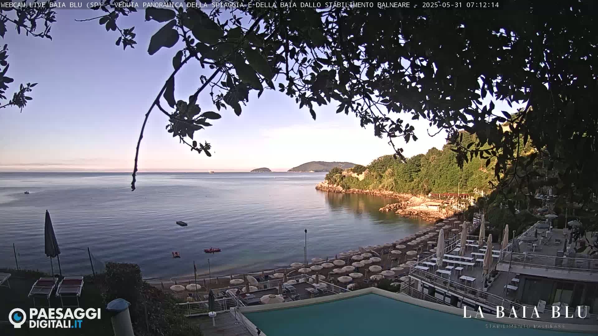 A calm bay with a sandy beach lined with umbrellas and outdoor seating is partially visible through tree branches, under a clear, light-blue sky.
