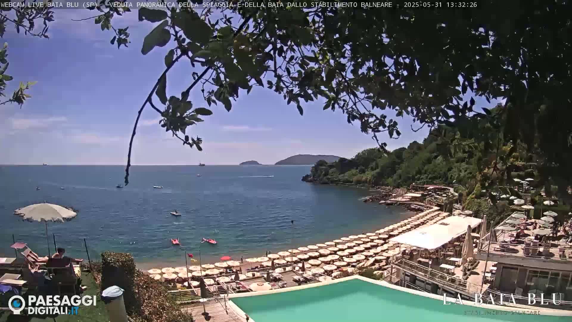 A sunny beach scene shows a calm bay with a sandy beach lined with rows of umbrellas and lounge chairs, an infinity pool, and green hills in the background.
