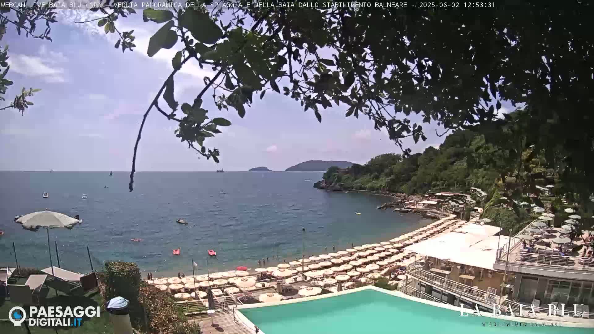 A partly sunny view from a hillside overlooking a calm bay with a beach full of umbrellas and a swimming pool, partially obscured by tree branches.