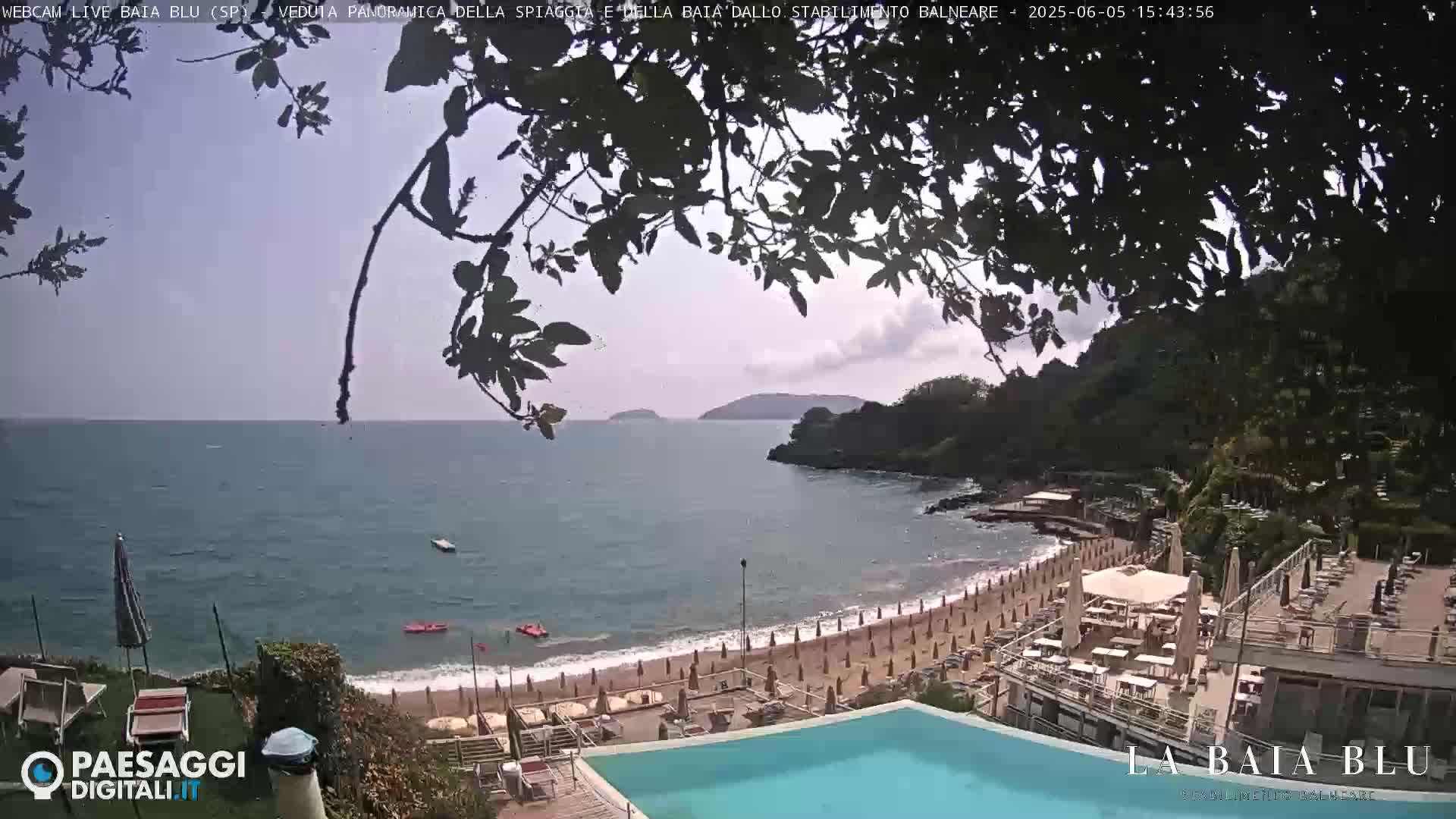 An overlook of a sandy beach with umbrellas and lounge chairs, a swimming pool, and calm blue water under a partly cloudy sky.